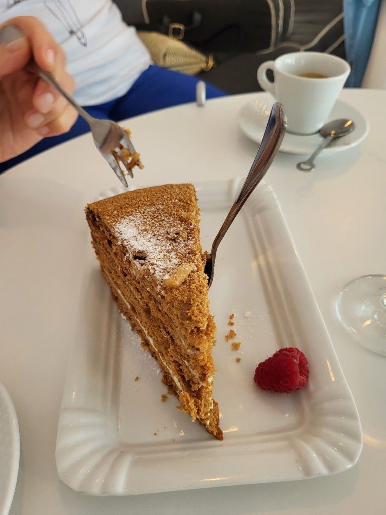 Portrait photo of a rectangular white plate holding a slice of Honey Nut cake dusted with icing sugar and with a single raspberry beside it. A small cake fork sticks out of the cake, and another fork hovers menacingly over the cake, ready to demolish it. In the background we can see a white cafe table on which a cup of espresso rests.