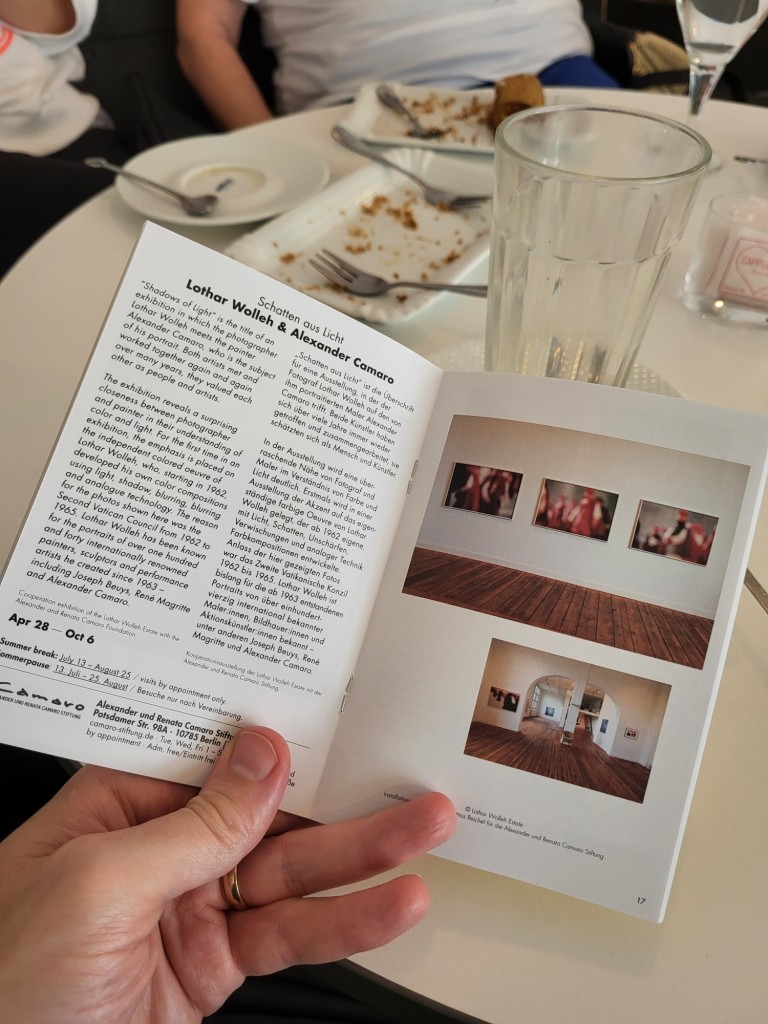 Portrait photo showing someone's hand holding open a copy of the Photography in Berlin A6 zine. The page spread displayed shows information about a photography exhibition by Lothar Wolleh & Alexander Camaro and some photos from the exhibition. In the background we can see a cafe table with plates bereft of cake, and glasses no longer containing the caffeinated beverages they once held.